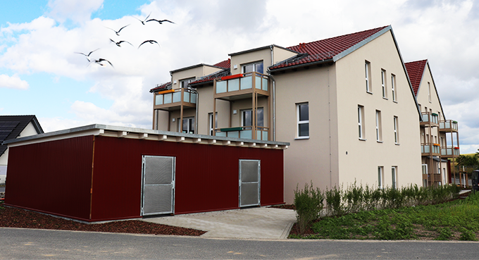 Ansicht des Außenbereichs der Tagespflegeeinrichtung in Arnsdorf bei Dresden. Der gepflegte Garten mit bunten Blumenbeeten und einem gemütlichen Sitzbereich lädt zu entspannenden Momenten im Freien ein. Rund um das Gebäude erstreckt sich eine grüne Wiese, die von Bäumen gesäumt wird und eine ruhige, naturverbundene Atmosphäre bietet. Der Eingangsbereich ist barrierefrei zugänglich und sorgt für einen einladenden Eindruck.